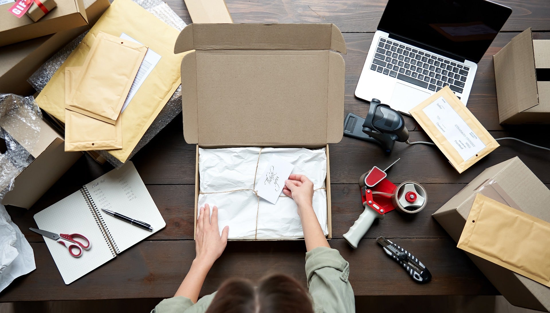 woman packing corrugated shipping box with tissue paper on table next to laptop and packing tape